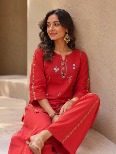 Woman wearing a vibrant red embroidered kurta and pants set with gold accents, sitting casually.