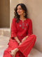 Woman wearing a vibrant red embroidered kurta and pants set with gold accents, sitting casually.