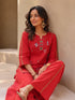 Woman wearing a vibrant red embroidered kurta and pants set with gold accents, sitting casually.
