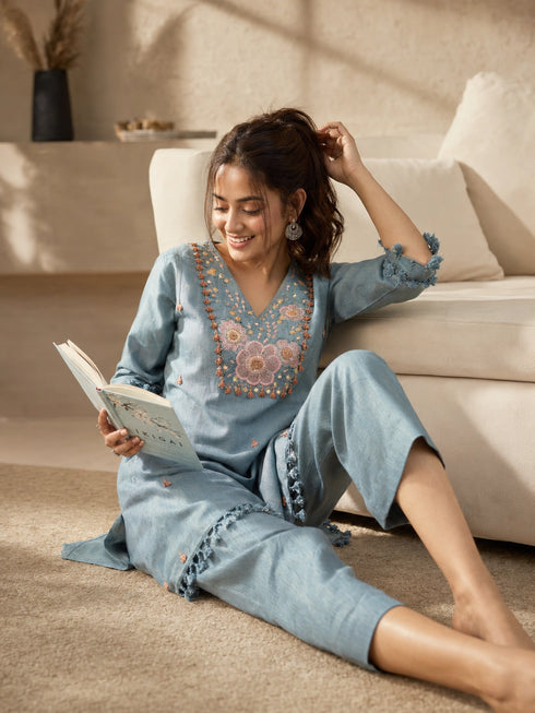Woman wearing a blue linen co-ord set with floral embroidery and tassel details, sitting and reading a book.