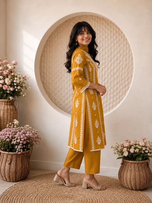 Woman wearing a mustard yellow embroidered kurta and pants set, sitting indoors with flowers.
