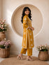 Woman wearing a mustard yellow embroidered kurta and pants set, sitting indoors with flowers.