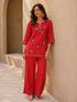 Woman wearing a vibrant red embroidered kurta and pants set with gold accents, sitting casually.