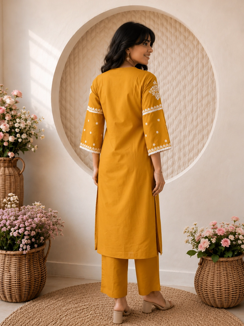 Woman wearing a mustard yellow embroidered kurta and pants set, sitting indoors with flowers.
