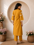Woman wearing a mustard yellow embroidered kurta and pants set, sitting indoors with flowers.