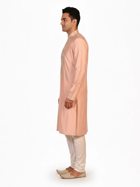 Man wearing a peach silk Kurta Pajama with intricate embroidery on the neckline and cream churidar pants.