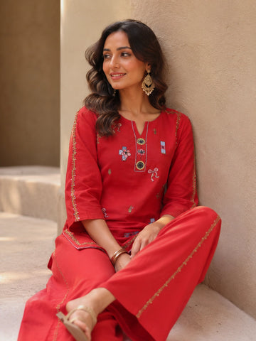 Woman wearing a vibrant red embroidered kurta and pants set with gold accents, sitting casually.