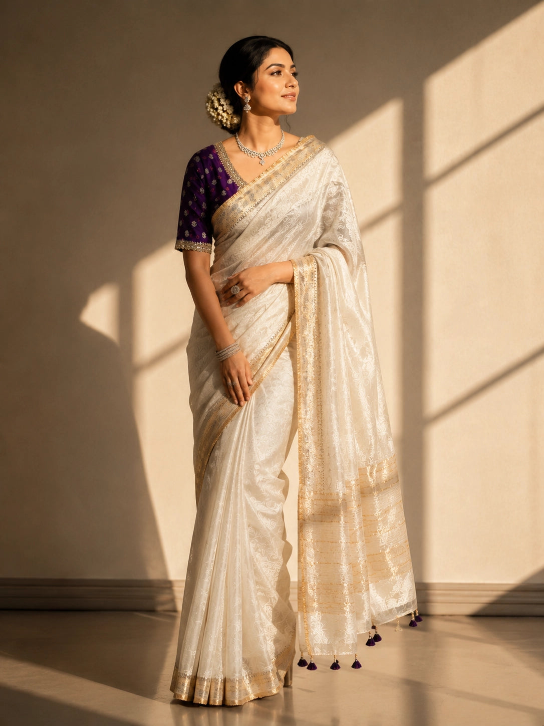 Woman in luxurious cream saree with gold border and embroidered purple blouse.