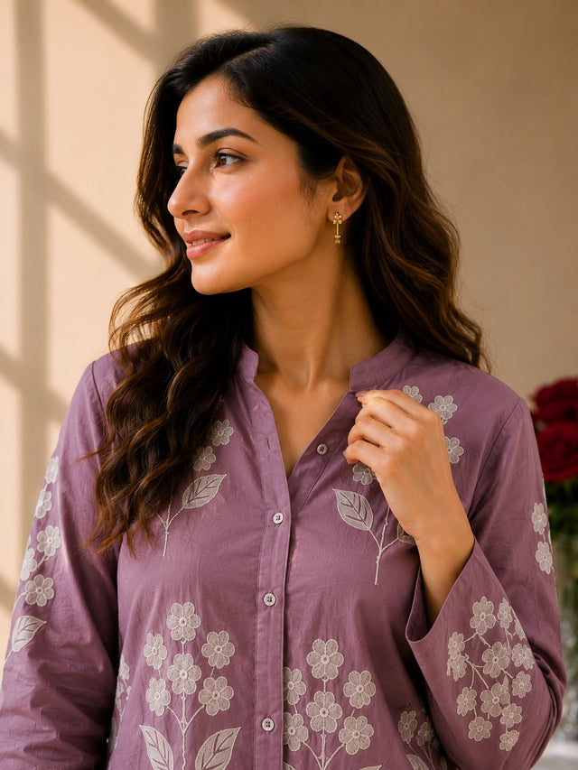 Smiling woman in a purple tunic with white floral embroidery and white pants.