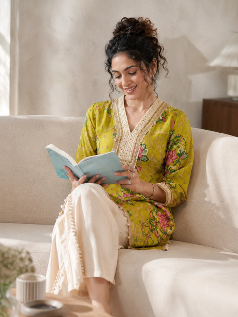Woman wearing a yellow floral print kurta with cream pom-pom pants, sitting on a sofa and reading a book.