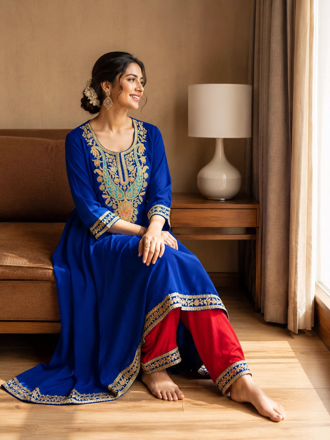 Woman in royal blue embroidered kurta with red pants, sitting by window.