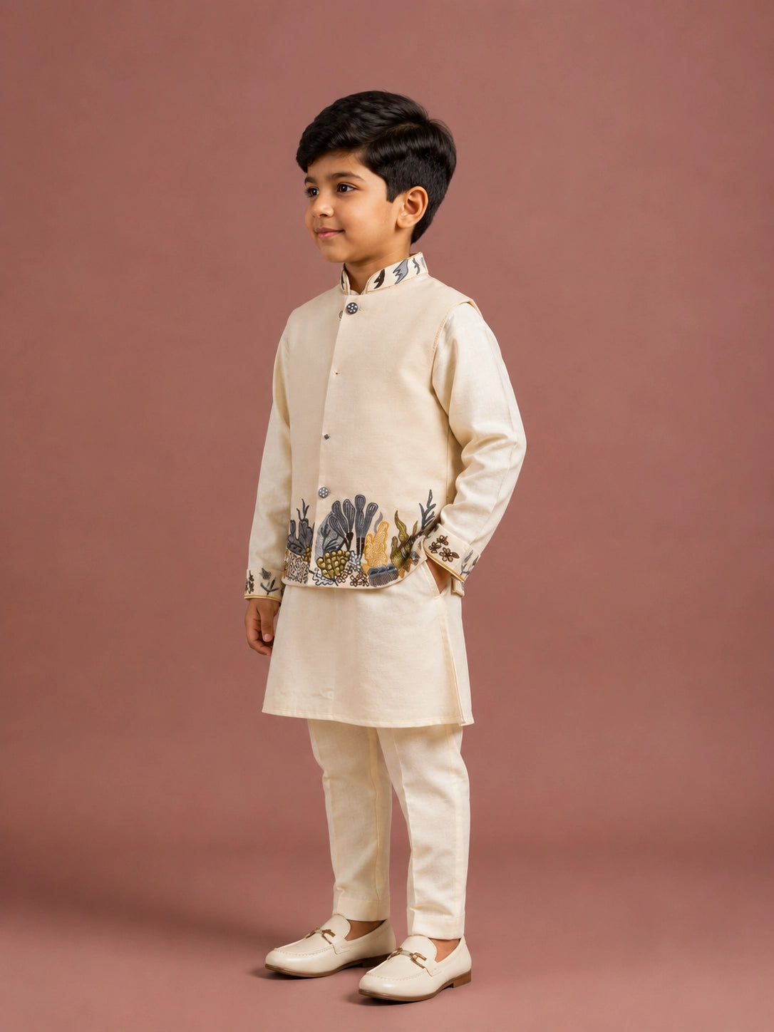 Young boy in a cream embroidered kurta and jacket set, sitting elegantly.