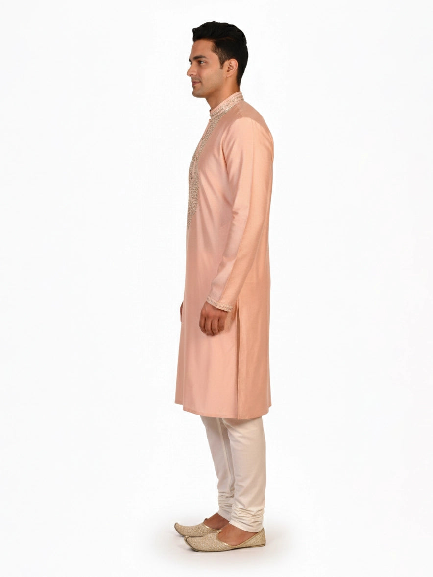 Man wearing a peach silk Kurta Pajama with intricate embroidery on the neckline and cream churidar pants.